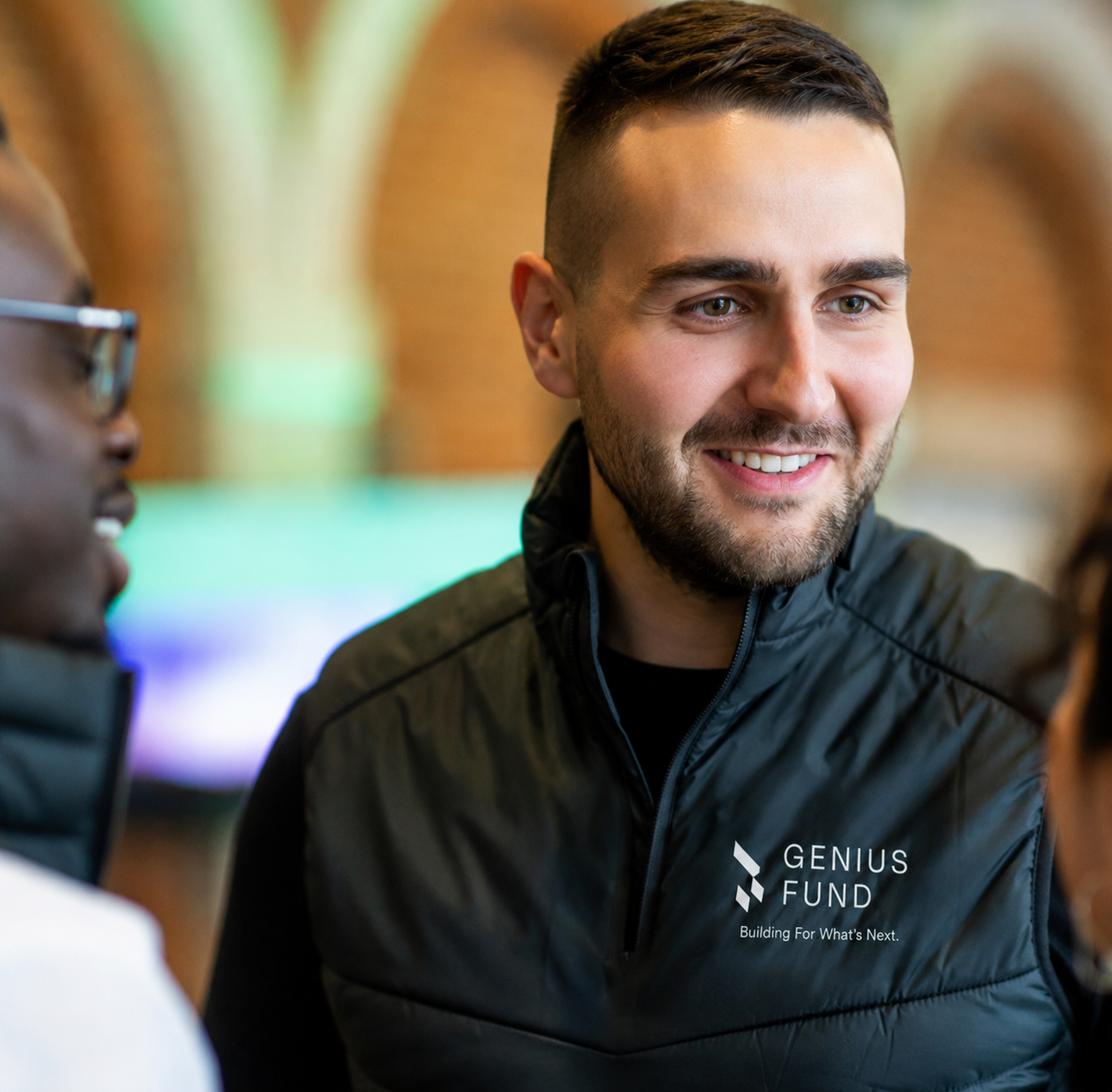 A Genius Fund team member in a branded vest, mid-conversation