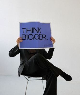 A person seated holding a large sign reading Think Bigger